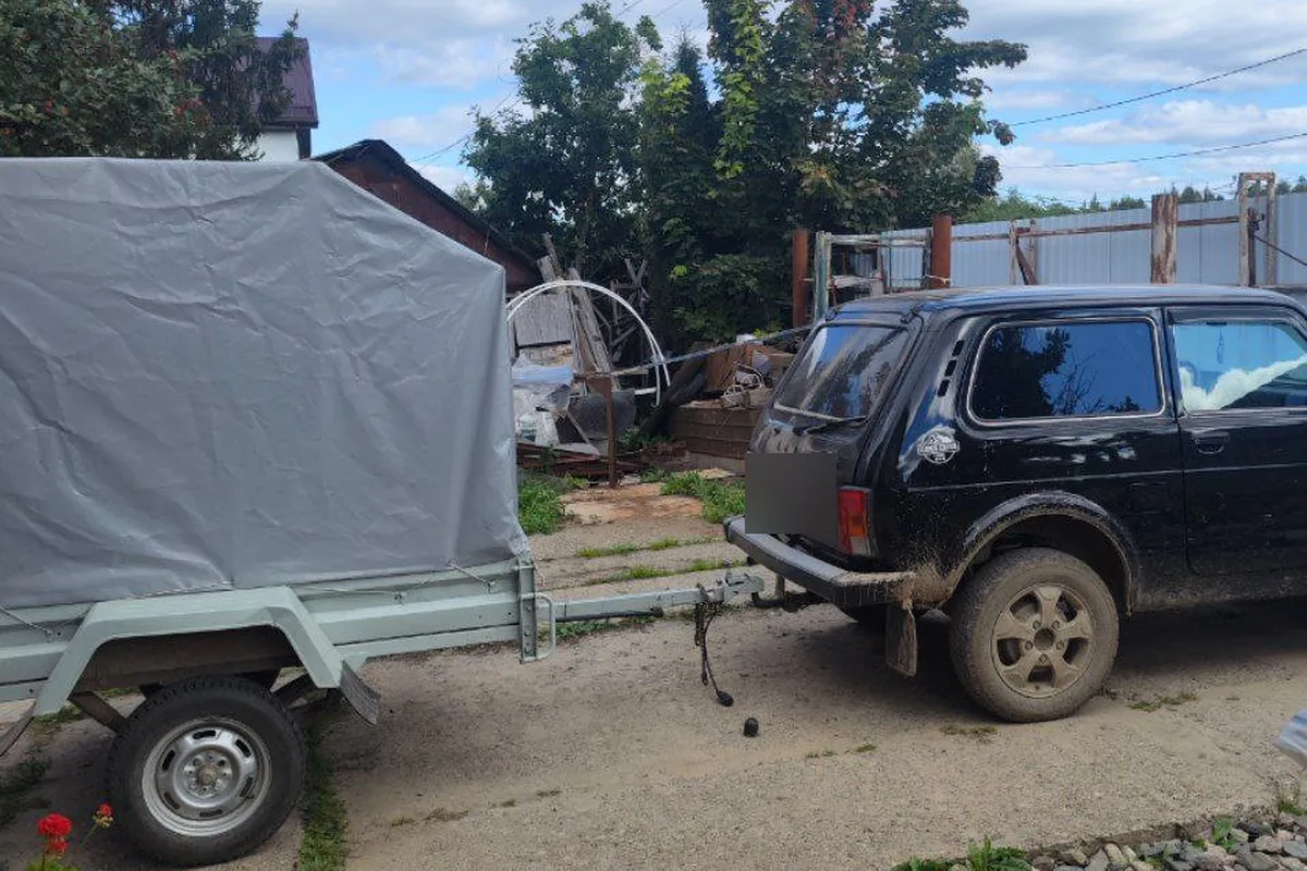 Чёрная пятидверная Нива с одноосным прицепом под тентом во дворе
